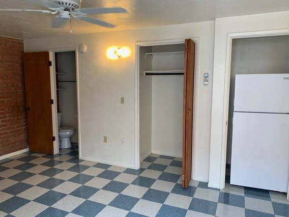 Interior view of a small studio apartment showing a checkered blue-and-white tile floor, an open closet with folding doors, a bathroom doorway with a visible toilet, and a white refrigerator in an alcove. A ceiling fan and wall light fixture are mounted above, and the walls are a mix of white paint and exposed brick.