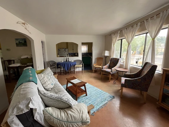 A cozy living room with large windows and white curtains, furnished with a cushioned sofa, two patterned armchairs, a small wooden coffee table on a blue rug, and a dining table covered with a blue cloth near an arched doorway leading to the kitchen.