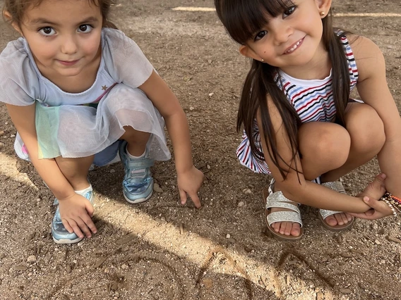 Two young children crouch on a dirt playground, smiling and pointing to letters they’ve drawn in the soil. One wears a light dress and sneakers, and the other wears a striped dress and sandals.