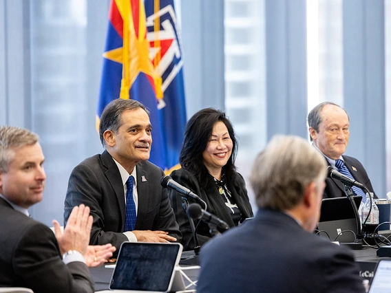 Several men and women take part in a discussion near the Arizona state flag and the University of Arizona flag