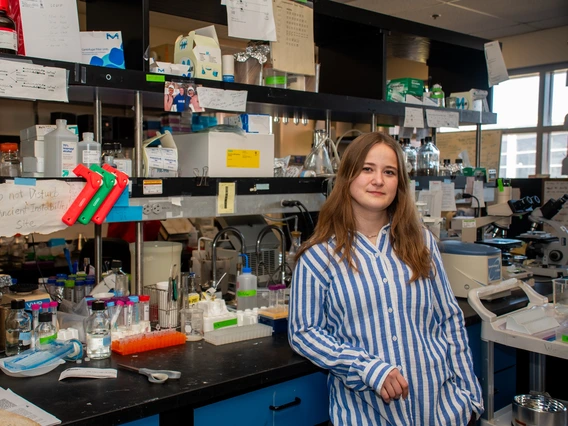 A laboratory setting with a young woman standing confidently in the foreground, in front of a cluttered workstation. The woman wears a blue and white striped shirt and has long brown hair. The laboratory bench is filled with various scientific equipment, including bottles, pipettes, and test tube racks.