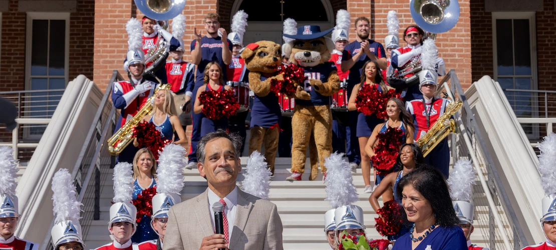 suresh garimella speaks in front of a building with a marching band and wildcat mascots behind him