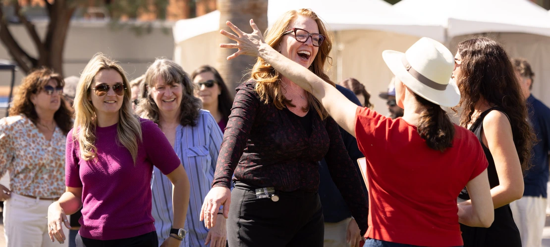 A group of smiling staff members gather outdoors at a social event. In the foreground, two women are sharing a joyful moment, with one reaching out to greet the other. The scene captures several others in the background, walking and chatting. The setting is sunny with white tents visible in the background.
