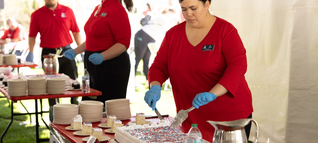 Two staff members in red shirts serve cake at a social event under a tent. One woman is focused on cutting a sheet cake with blue gloves, while the other, smiling, stands in the background. Plates with cake slices are arranged on the table, and there are several empty plates and a water bottle nearby. The event appears lively and well-organized.