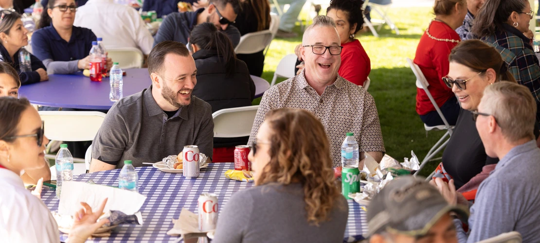 A group of staff members enjoy lunch at a social event under a tent. Several people are seated around a table, smiling and laughing while chatting. Two men in the foreground, one in a grey shirt and the other in a patterned shirt, are laughing together. Bottles of water, sodas, and food wrappers are visible on the table. In the background, other staff members are seated and enjoying the event.