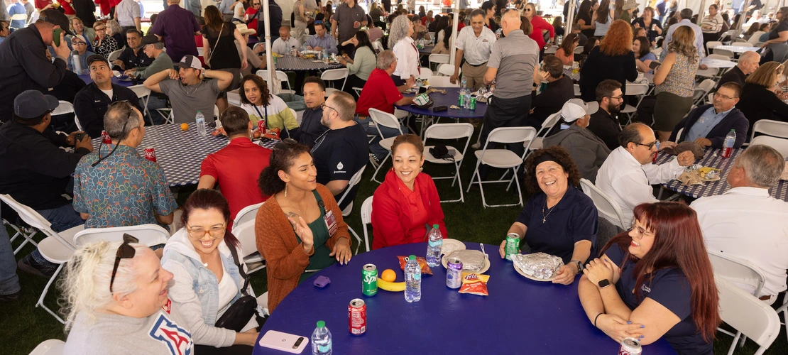 Several staff members sit around tables under a large tent at a social event. In the foreground, a group of colleagues smiles and enjoys their meal, with drinks, snacks, and wrapped food on the table. The people are wearing a mix of casual clothing, and the atmosphere is lively, with more staff members visible in the background, socializing and eating.