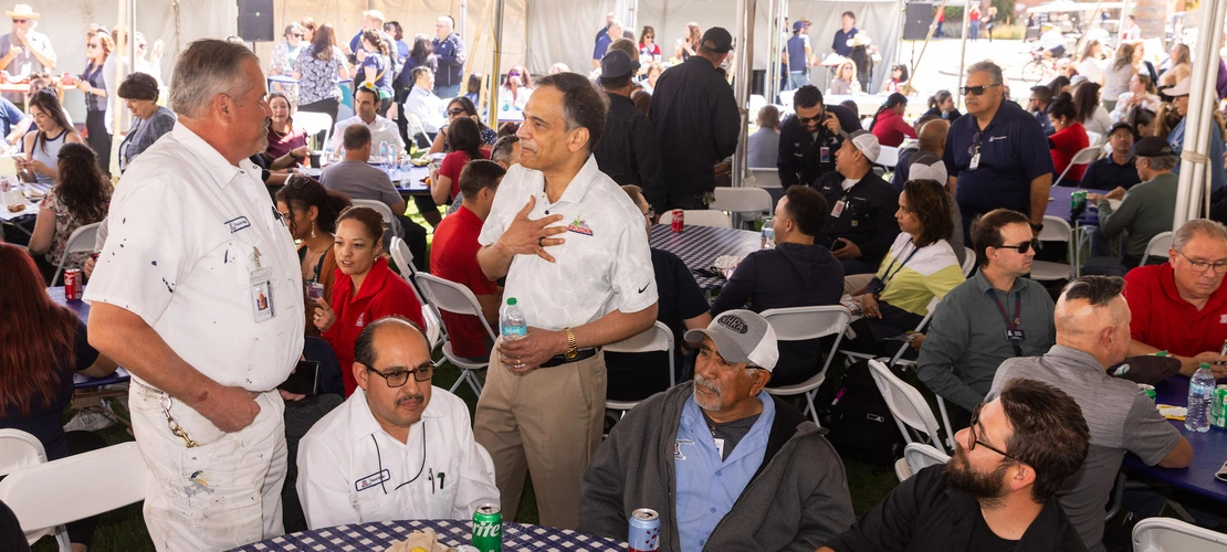 University of Arizona President Suresh Garimella stands and chats with staff members at a social event under a large tent. One staff member, dressed in a white shirt, is speaking to Garimella while others sit at a table nearby, enjoying their meal. The scene is lively with groups of people engaging in conversation, eating, and socializing. Several staff members are visible in the background, creating a relaxed and friendly atmosphere.
