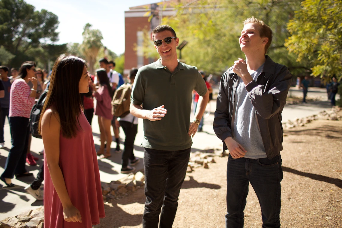 Three University of Arizona students stand and chat in a sunny outdoor area near Old Main. The student on the left wears a sleeveless pink dress, the student in the middle wears sunglasses and a dark green polo shirt, and the student on the right wears a gray T-shirt and jacket. Other students can be seen talking and walking in the background beneath tall trees.