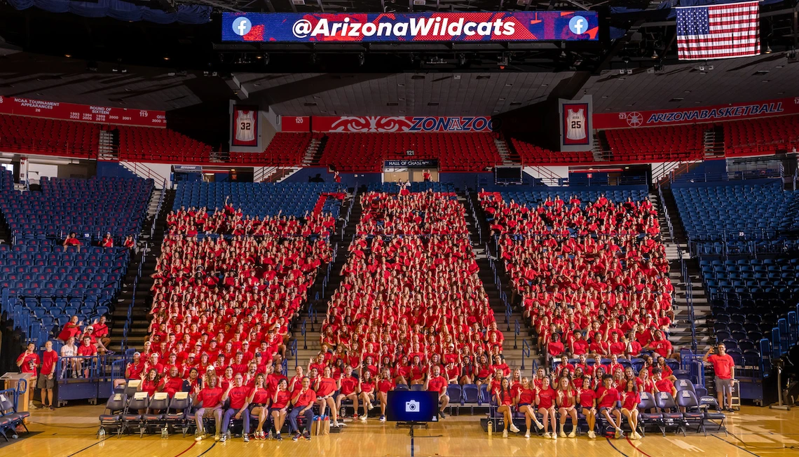 Hundreds of University of Arizona student-athletes wearing red shirts sit together in the shape of a large block “A” inside McKale Memorial Center during the Arizona Athletics welcome back BBQ. An American flag hangs above, and a scoreboard displays the @ArizonaWildcats handle.