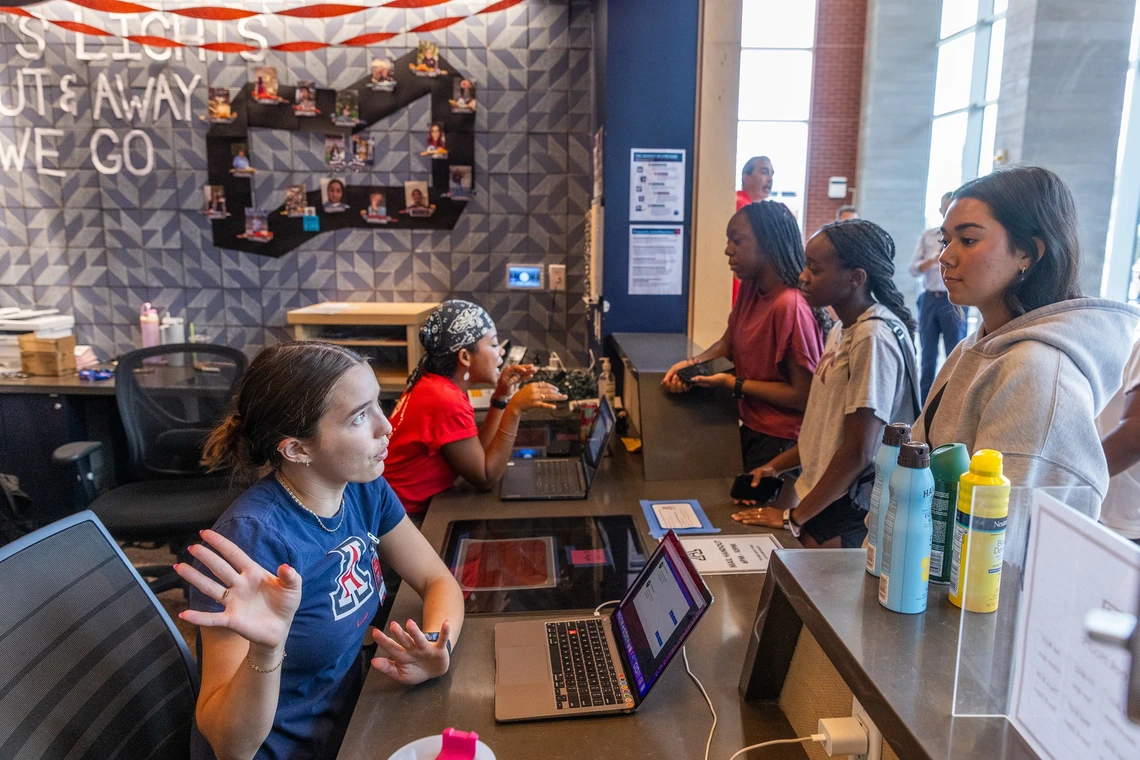 A pair of women wearing University of Arizona branded t-shirts sit the front desk of a student housing building. Standing in front of the desk are three female students wearing casual clothing.