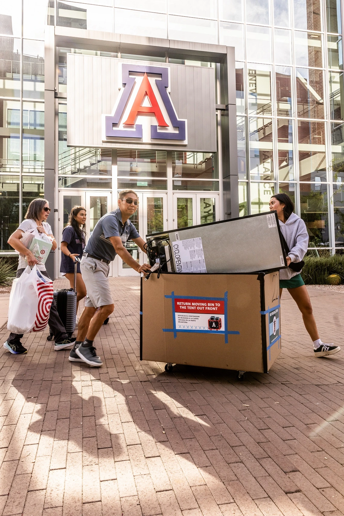 A middle aged man and woman help a pair of students push a large cardboard cart full of bedroom apparel and supplies into a college dorm. A large, glass-front building is in the background, with a large block "A" logo at the top.