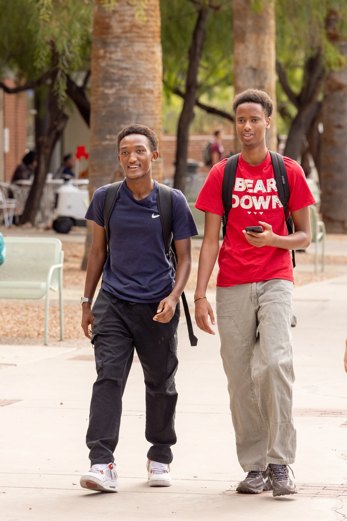 A man wearing black pants and a blue polo shirt and backpack walks next to a man wearing tan slacks and a red t-shirt that says "Bear Down." The two men are walking on a university campus with trees and seating in the background.