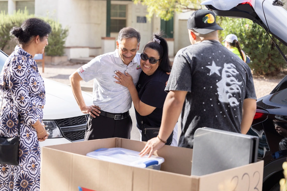 A man wearing a white, long sleeve button down greets a family outside of their vehicle. There is a large cardboard cart in front of the group containing a collection of dorm room decorations and supplies.