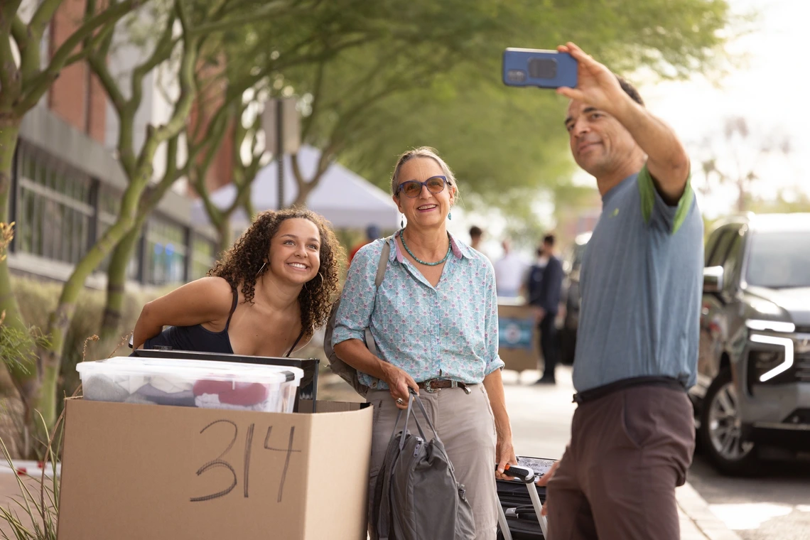 A young woman with curly brown hair poses for a selfie with an older woman wearing a light blue button down and grey slacks and a man wearing jeans and a blue t shirt. They are standing outside of a brick-built, college dorm building.
