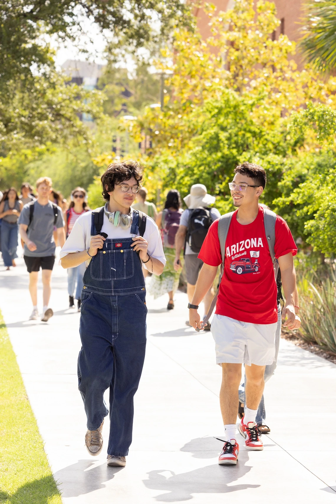 A man wearing overalls and a white t-shirt walks next to a man wearing white shorts and a red t-shirt, along a sidewalk on a university campus. There is grass, desert trees and other students in the background.