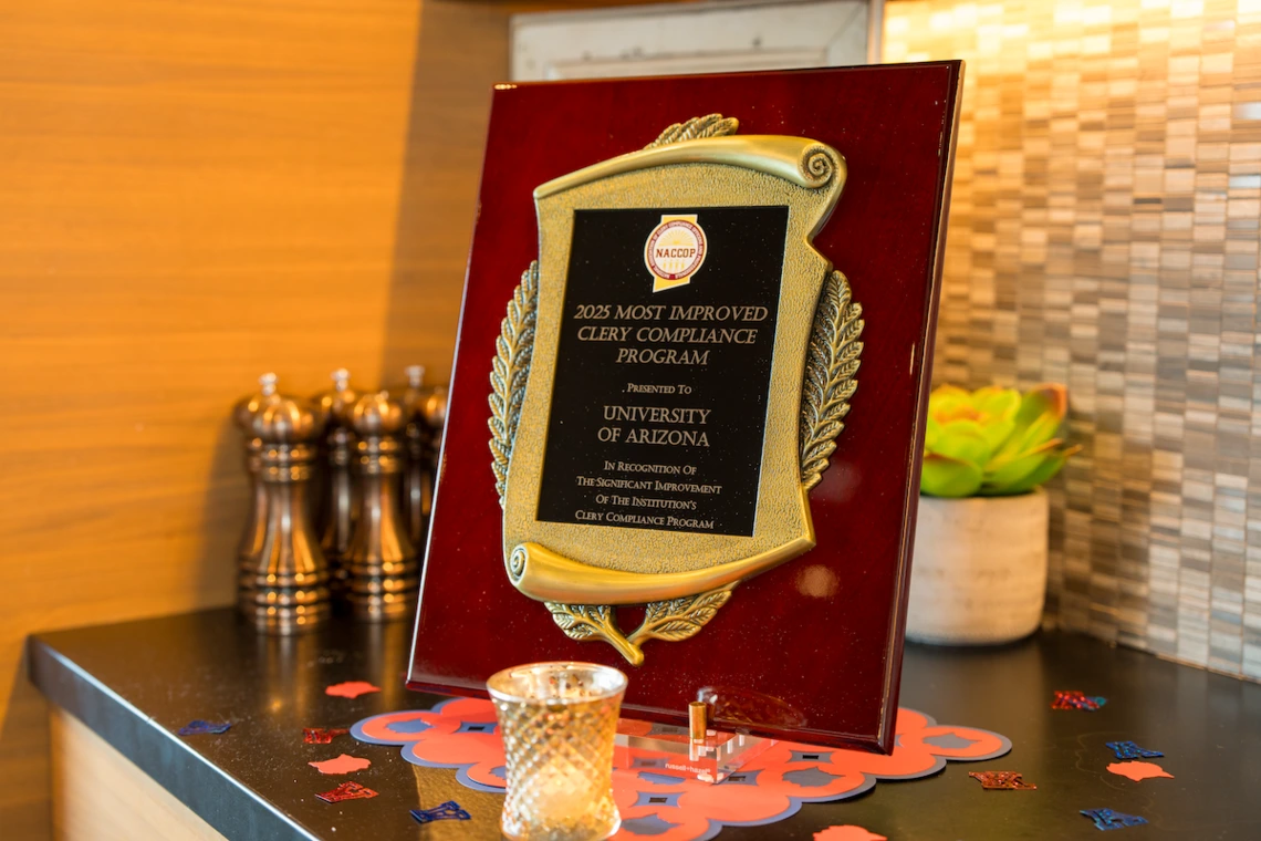 A close-up of a plaque reading "2025 Most Improved Clery Compliance Program, presented to University of Arizona," displayed on a countertop with small decorative items around it.