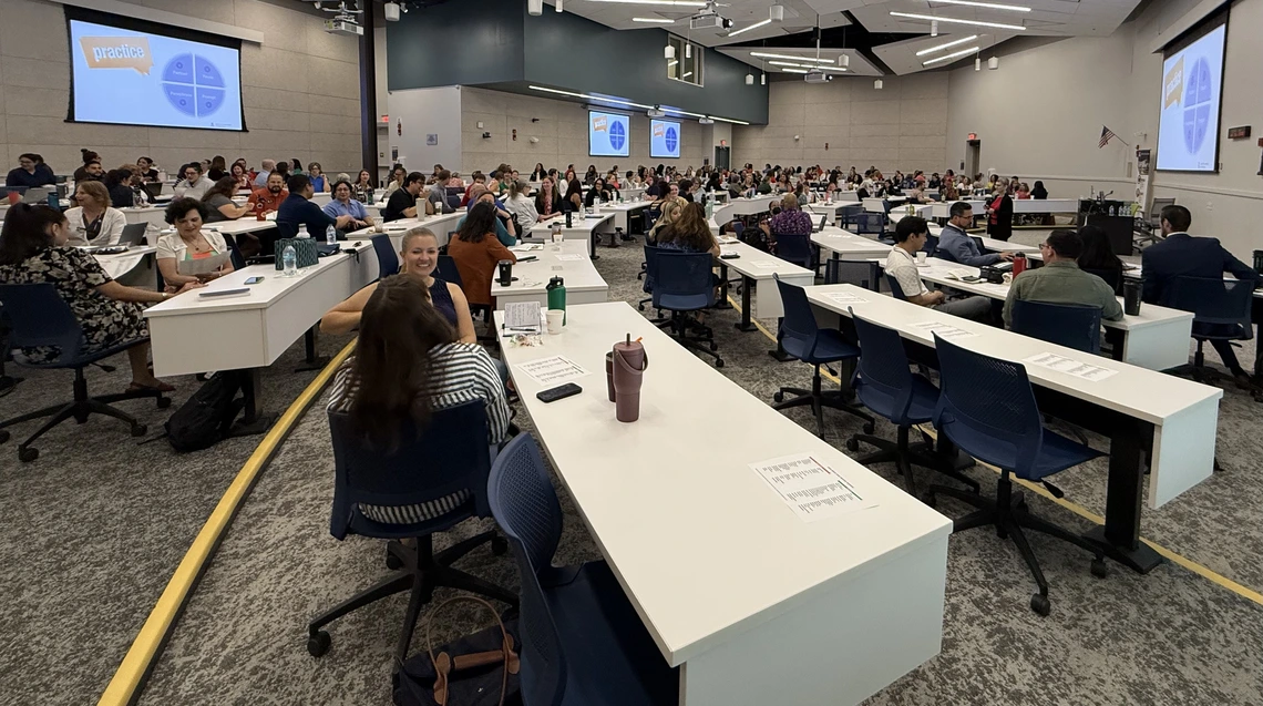 Attendees fill a large lecture hall during the July 30 Conflict Resolution and De-escalation Skills Symposium at the University of Arizona. Participants sit in groups at long white tables, engaging in discussion during an interactive portion of the event. Multiple presentation screens display a slide with the word “practice” and a blue circular graphic divided into four sections.