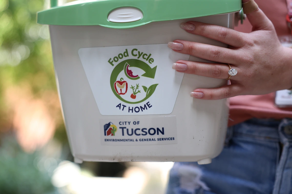 A close-up of a person holding a small white composting bin labeled “Food Cycle at Home,” with logos for the City of Tucson Environmental & General Services. The person’s hand, wearing a large oval-cut engagement ring, rests on the bin.