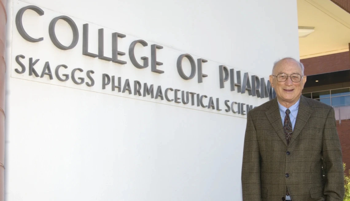Jack R. Cole, wearing a brown plaid suit and patterned tie, stands smiling in front of a white wall with large lettering that reads "College of Pharmacy Skaggs Pharmaceutical Sciences."