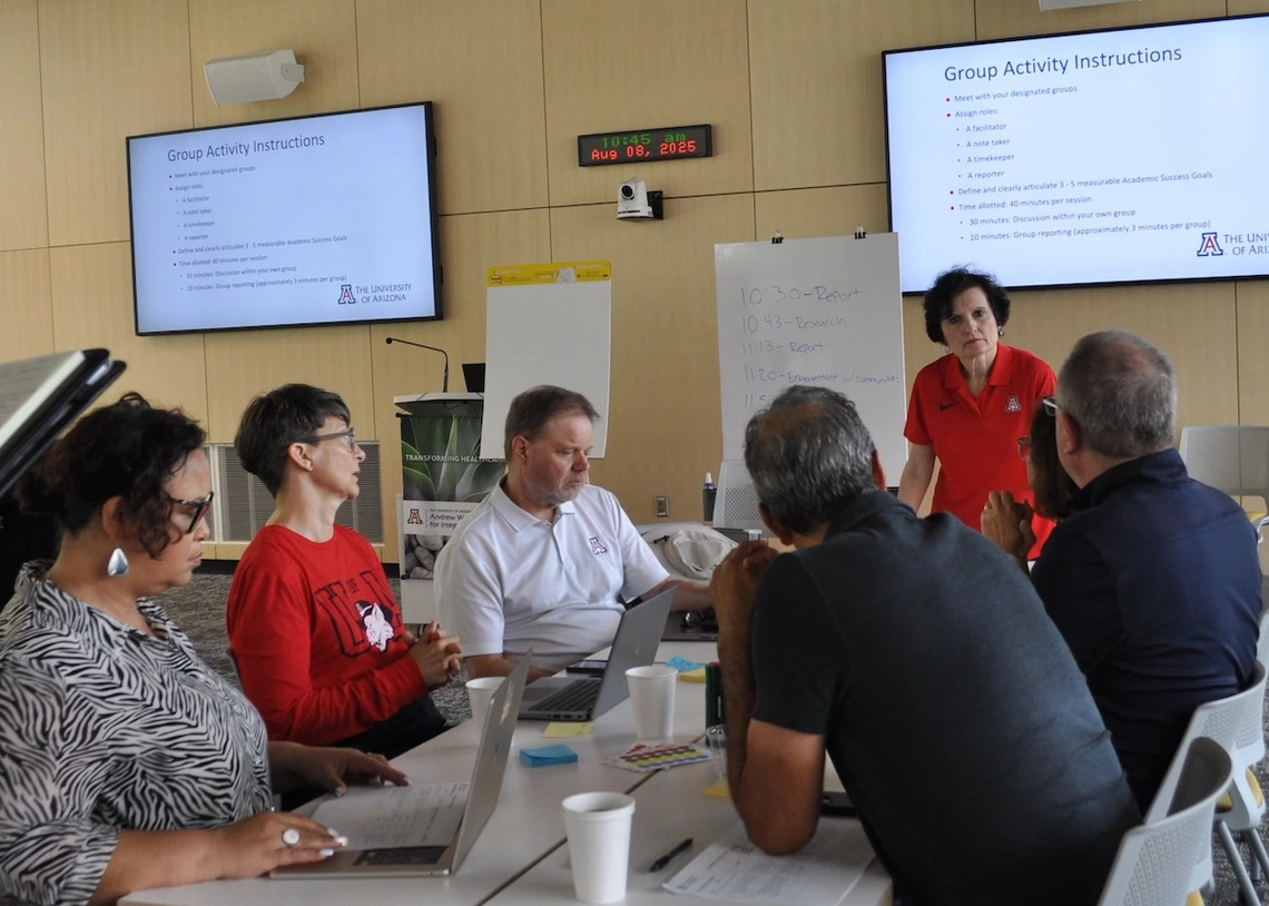  A group of six people sit around a table in a meeting room at the University of Arizona, listening to a woman in a red U of A polo shirt who is speaking to them. Laptops, papers, and coffee cups are on the table, and two screens behind them display “Group Activity Instructions.”