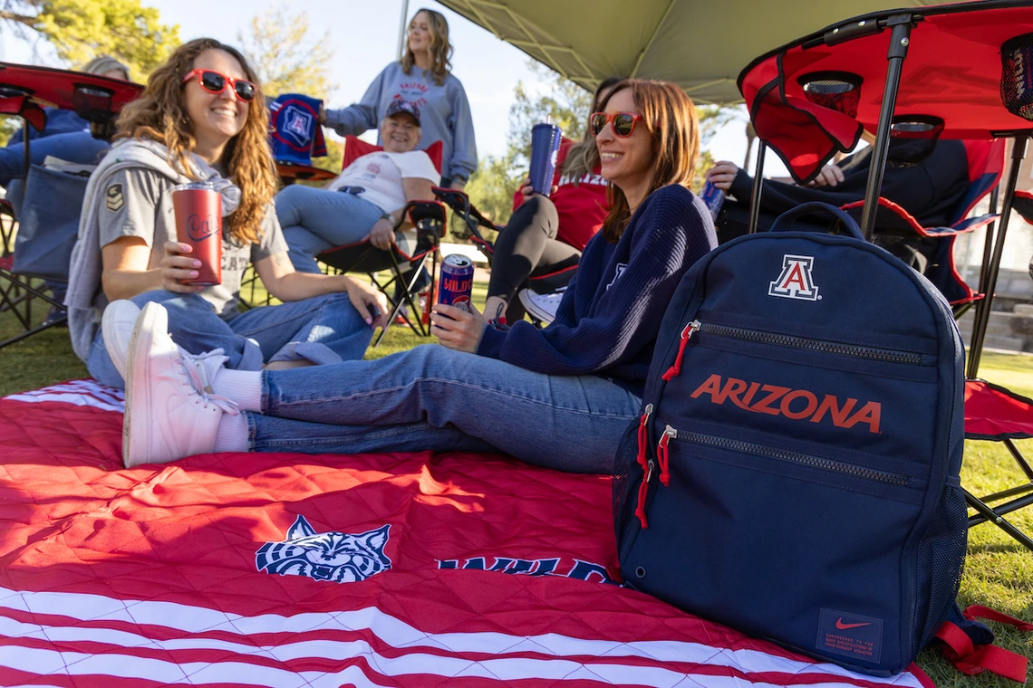 People relax under a canopy at a tailgate on a sunny day, sitting on a red University of Arizona blanket. A navy backpack with “Arizona” and the “A” logo is in the foreground.