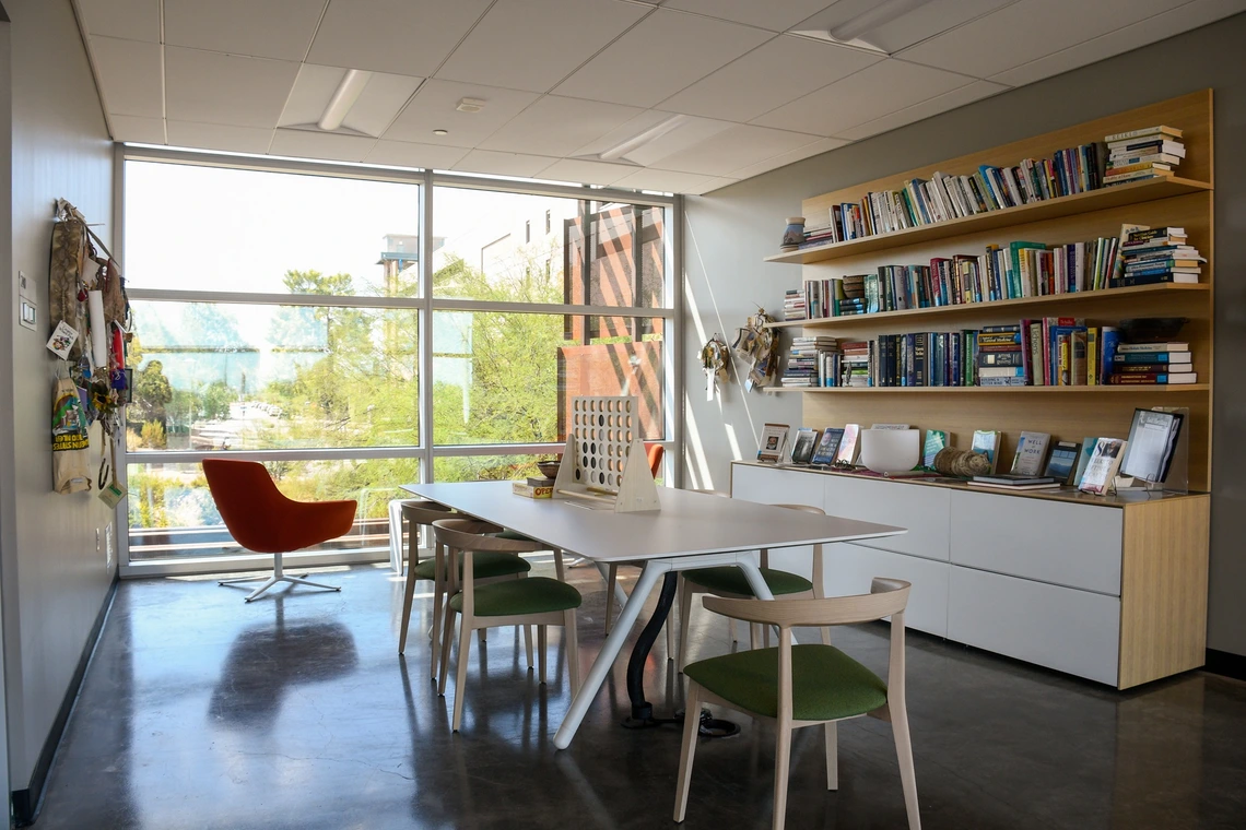 Modern office with large window, white table and chairs, orange lounge chair, bookshelves filled with books, and cabinets along the wall. Natural light fills the room.