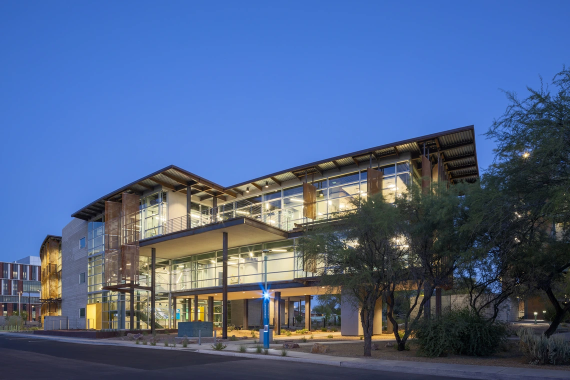 Modern three-story building with large glass windows, lit from inside at dusk, surrounded by trees and landscaping, with a blue emergency call box in front.