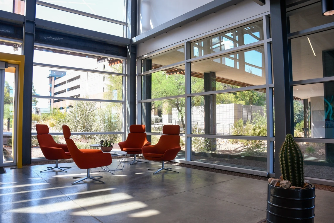 A modern indoor lounge area with floor-to-ceiling windows features four bright orange chairs arranged around a small table with a plant. Natural light fills the space, highlighting views of desert landscaping and nearby buildings outside, with a tall potted cactus in the foreground.
