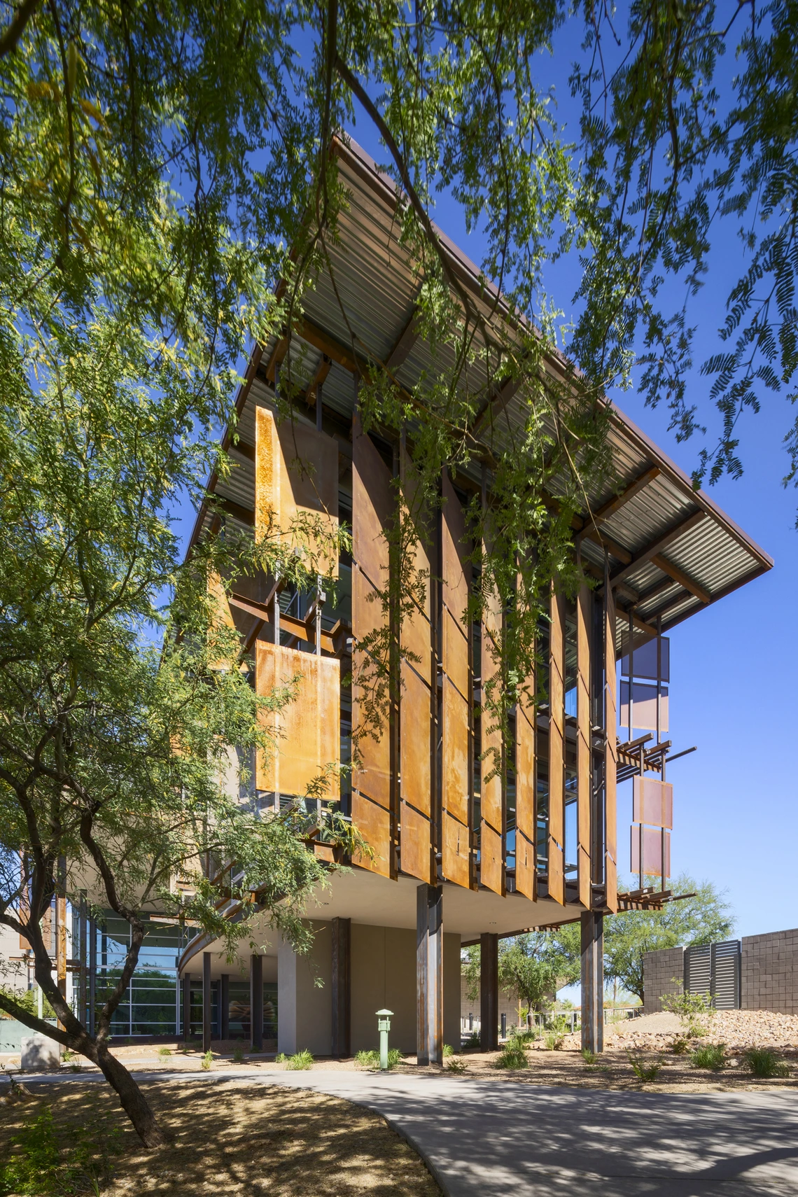 Modern building with tall rust-colored metal panels and large glass windows, elevated on stilts above a shaded walkway. Green trees frame the structure against a bright blue desert sky, blending natural and industrial design elements.