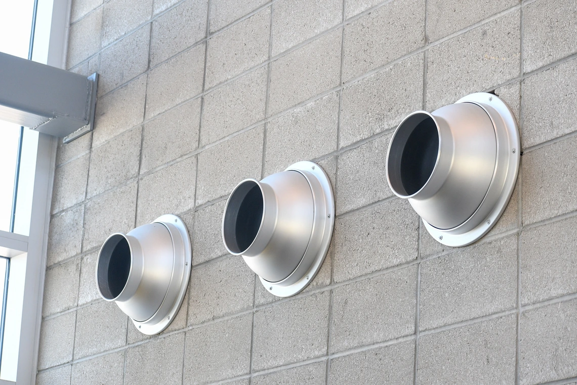 Close-up of three round metallic ventilation ducts mounted in a row on a gray concrete block wall. The vents project outward with a smooth, industrial design, catching light from nearby windows.
