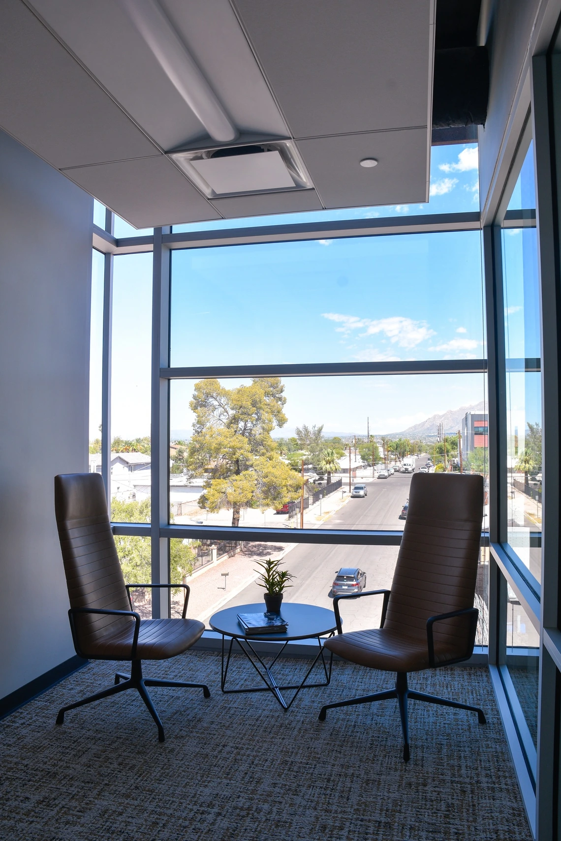 A modern office corner with floor-to-ceiling windows allowing a clear view of the street outside. Two brown leather chairs with metal frames are positioned on either side of a small round table. A small potted plant and a closed book or magazine rest on the table. Through the large windows, a sunny street scene is visible, featuring a wide road lined with trees and a few buildings. 