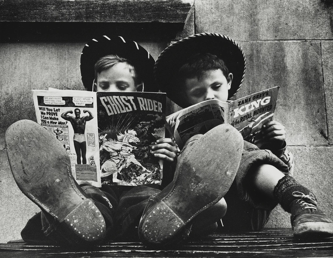 Two boys in cowboy hats sit side by side with their feet stretched out toward the camera, each reading a comic book. One holds The Ghost Rider while the other reads a Zane Grey western. The image is in black and white, highlighting their worn shoes and absorbed expressions.