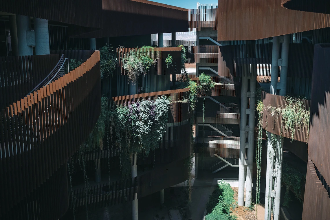 Interior view of the ENR2 Building’s canyon-like courtyard, with rust-colored fins, open-air walkways, and terraces covered in hanging vines and greenery.