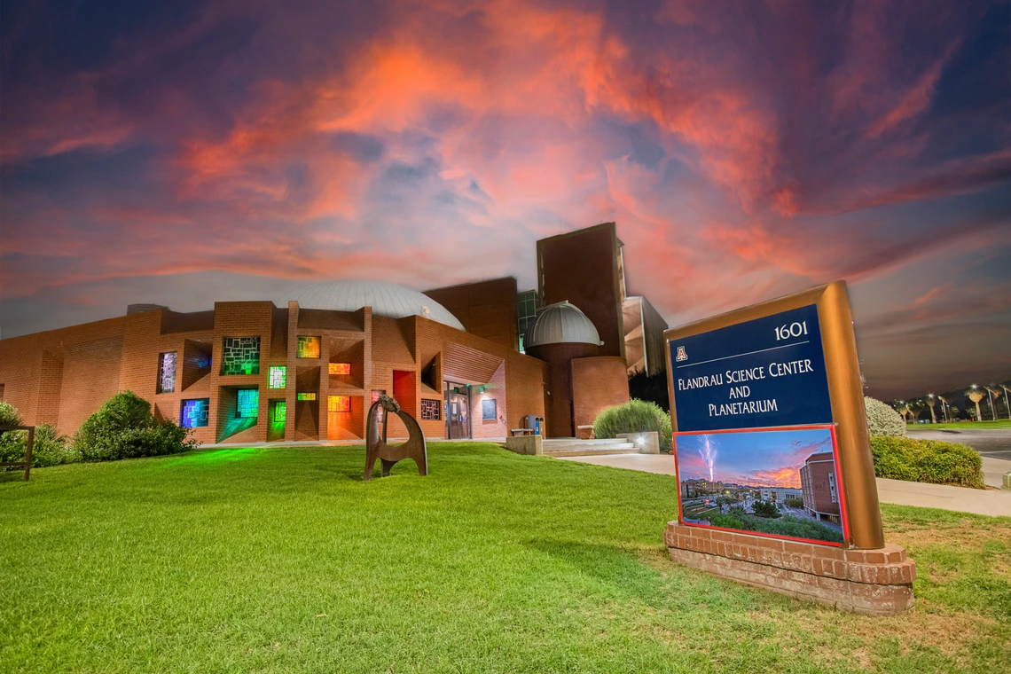 The exterior of the Flandrau Science Center and Planetarium at sunset. The building is constructed with red brick and features modern architectural designs, including a large dome that hints at its planetarium function. A series of rectangular windows are illuminated with various colors, creating a vibrant spectrum on the building's facade.