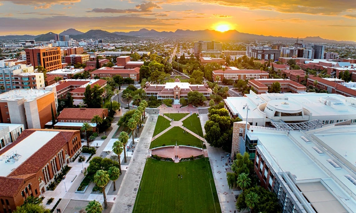 Aerial view of the University of Arizona campus at sunset, with Old Main at the center, the grassy Mall stretching toward the mountains in the distance, and the sky glowing with orange and purple clouds.