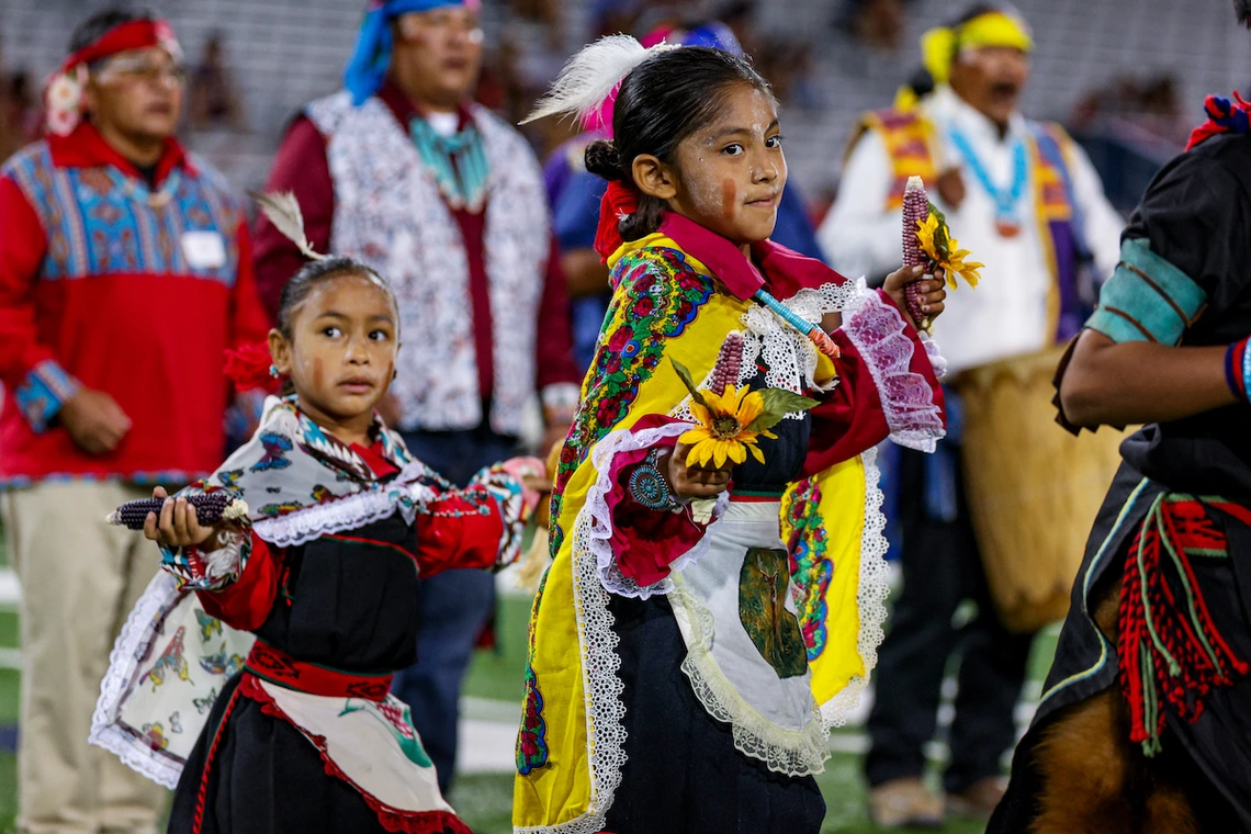 Two young children in traditional Hopi regalia perform a dance on the field, holding corn and flowers, during the Hopi Recognition Football Game at Arizona Stadium, with adult participants standing behind them.