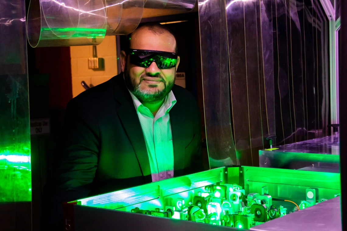 A man wearing safety glasses and a suit observes a green-lit optical experiment with lasers and mirrors in a laboratory setting.