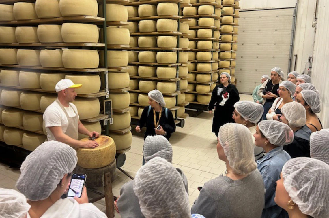 Students wearing hairnets gather around a worker in a cheese factory, listening as he explains the process in front of large wheels of cheese stacked high on shelves.