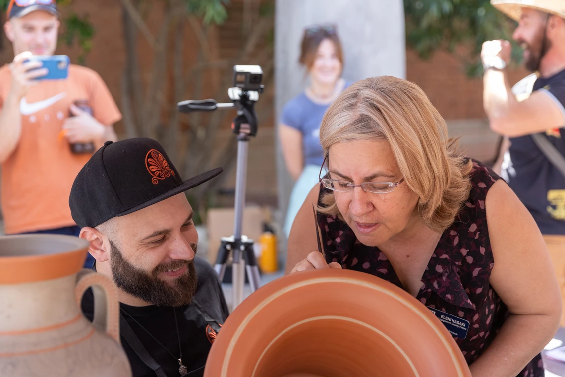 Eleni Hasaki looking over the shoulder of Roberto Paolini, an artist, to look at a reddish-colored vase that Paolini is painting a design on