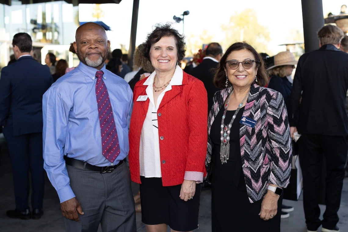 Three people stand together smiling at a reception outside the Stevie Eller Dance Theatre. The man on the left wears a blue shirt and red patterned tie, the woman in the center wears a red jacket, and the woman on the right wears a patterned jacket and glasses. Other guests mingle in the background.
