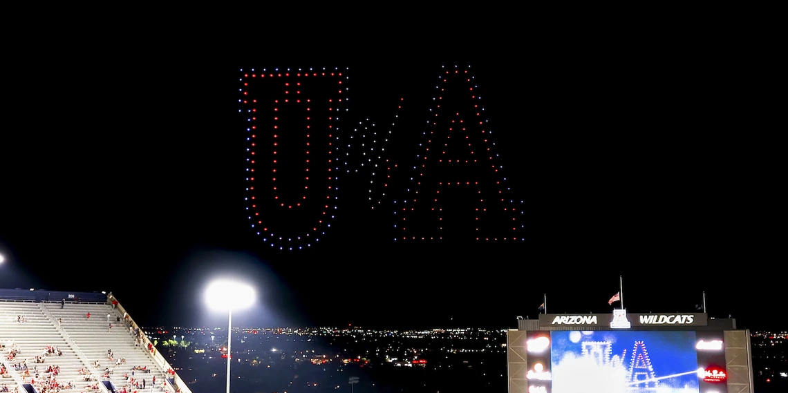 A nighttime drone show above Arizona Stadium forms a red, white, and blue "U of A" in the sky during a University of Arizona football game. The illuminated letters appear over the lit stands and scoreboard as the city lights of Tucson glow in the distance.
