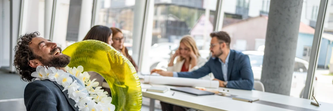A man wearing a flower lei and a large yellow pool float laughs while sitting at a conference table, as colleagues in business attire hold a meeting in the background. The playful scene contrasts the relaxed mood of the man with the focused group discussion in a bright, modern office.