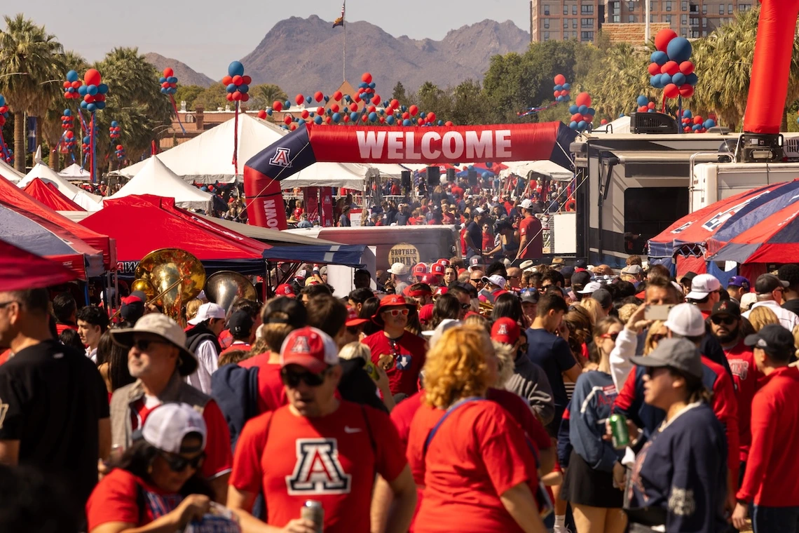 A large crowd of University of Arizona fans fills the University of Arizona Mall during Homecoming festivities, surrounded by red and blue tents, balloons, and a “Welcome” archway with mountains visible in the background.
