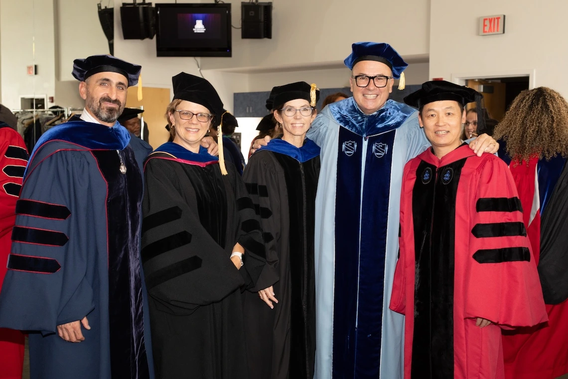 Five University of Arizona faculty members in academic regalia pose together and smile before a ceremony. They wear gowns in shades of blue, black, and red with velvet panels and caps, standing in a well-lit indoor space with other participants in the background.