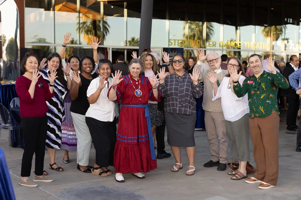 A group of people stand outside the Stevie Eller Dance Theatre, smiling and waving toward the camera. The group includes men and women in a variety of professional and traditional attire, gathered in front of the building’s glass entrance during a reception.