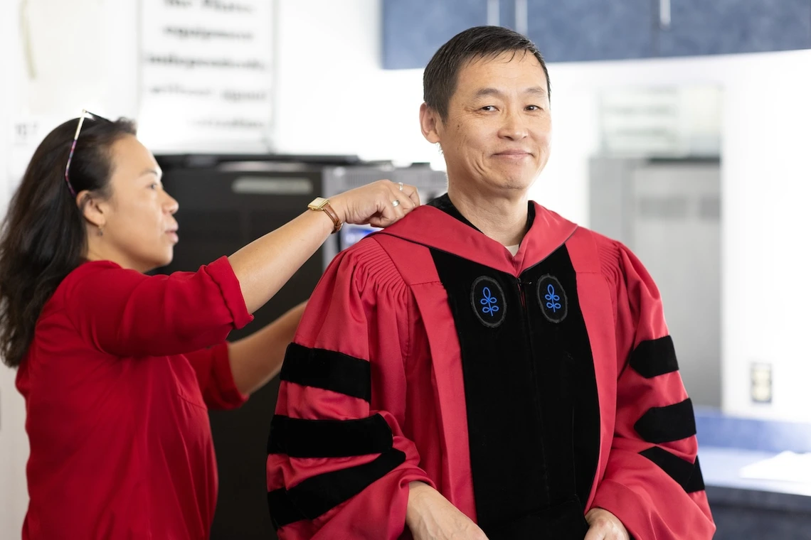 A woman helps a man adjust his academic regalia before a ceremony. The man, dressed in a red gown with black panels, smiles slightly while the woman, wearing a red top, focuses on the adjustment. The background shows an indoor space with soft lighting and blurred details.