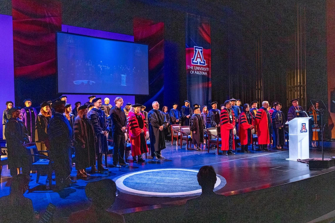 Faculty members in academic regalia stand on stage during the Luminaries Outstanding Faculty Awards ceremony at the University of Arizona. A large screen behind them shows a live feed of the event, and banners featuring the university’s logo hang on the stage.