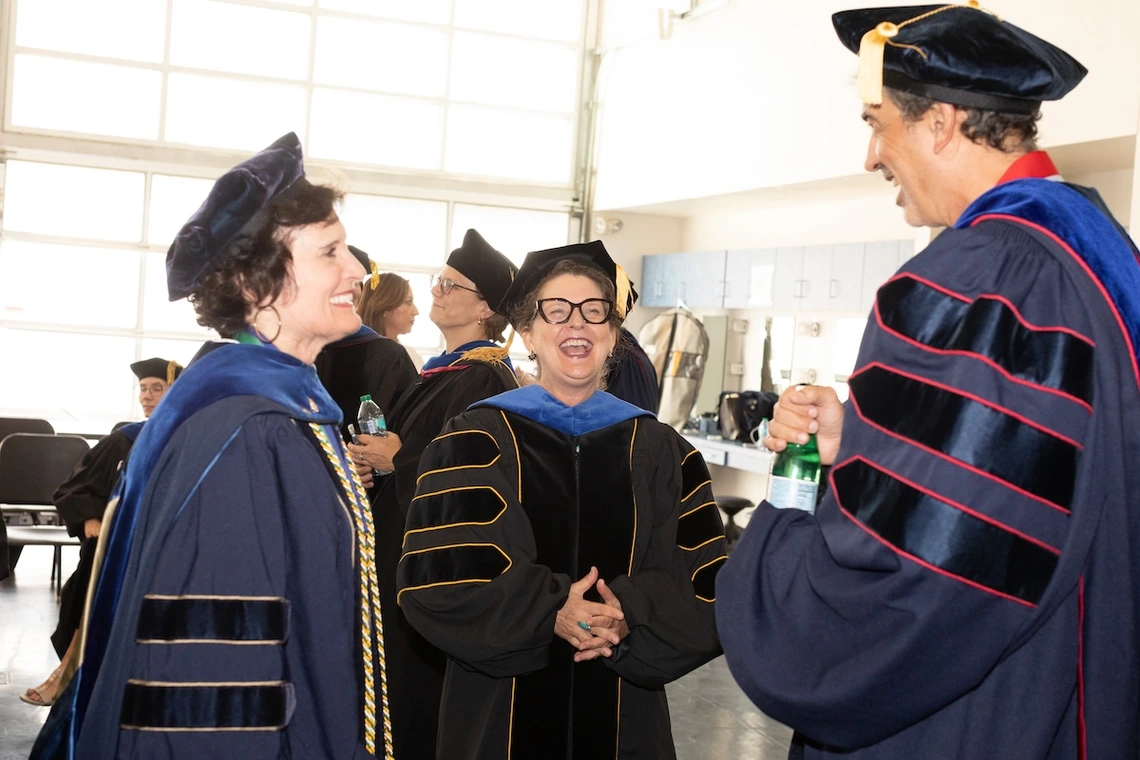 Three University of Arizona faculty members in academic regalia share a laugh before the Luminaries Outstanding Faculty Awards ceremony. Two hold water bottles as they chat in a brightly lit room with other participants in the background.