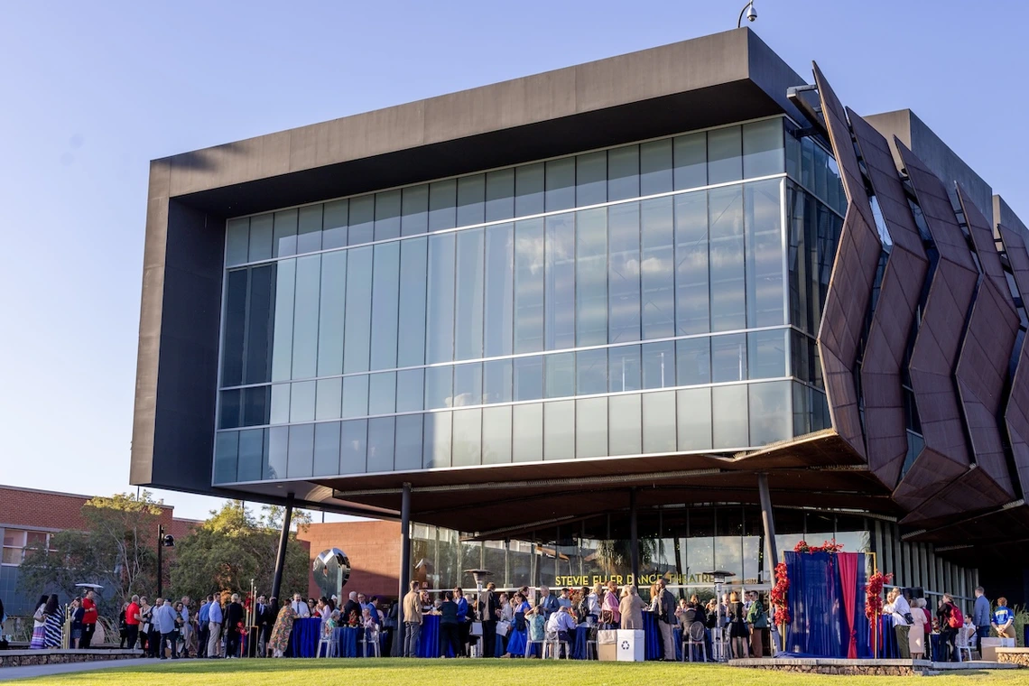 Guests gather for a reception outside the Stevie Eller Dance Theatre at the University of Arizona. People mingle around tables draped in blue cloths on the lawn beneath the building’s modern glass and metal façade on a clear afternoon.