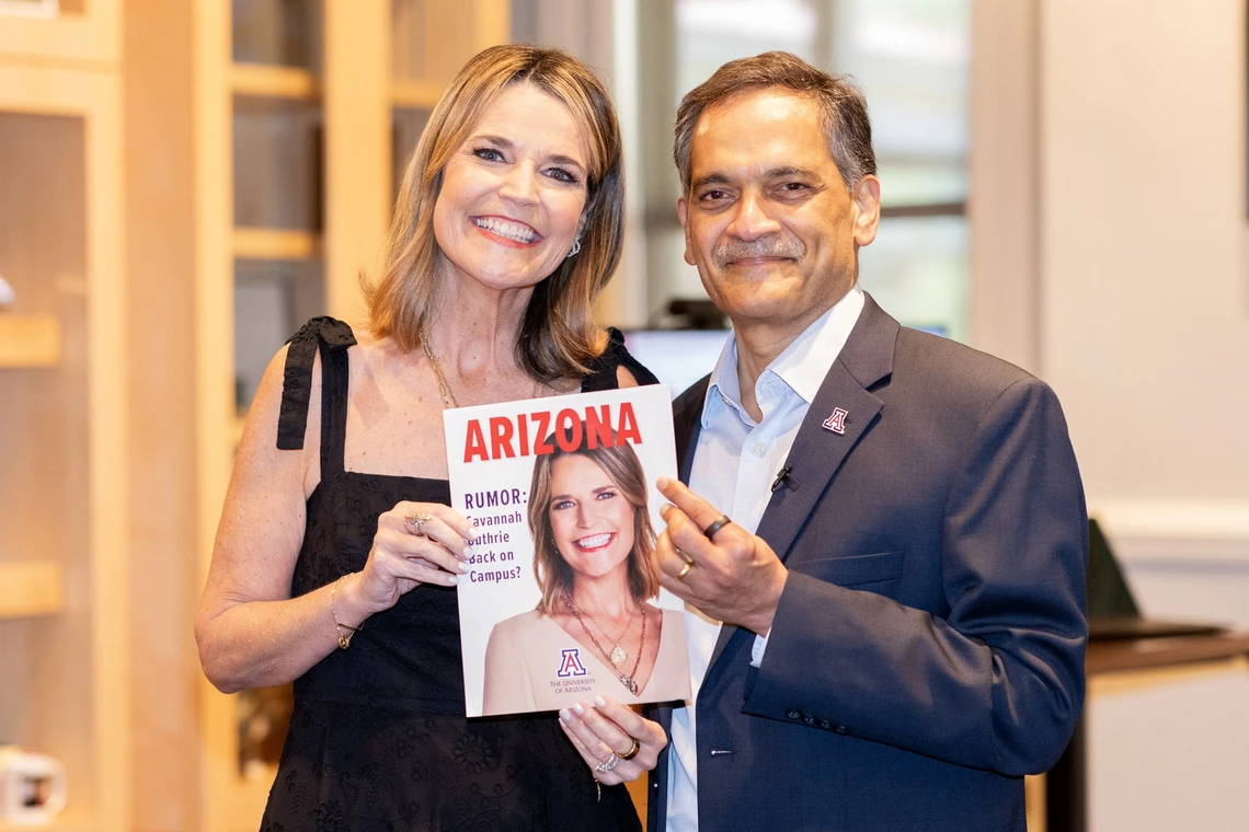Two individuals are posing indoors. The woman on the left is wearing a black sleeveless dress and a necklace. She is smiling broadly while holding a magazine with a cover that features her photograph. The man on the right is wearing a dark suit with a light-colored shirt and is smiling while pointing at the magazine. The background consists of a softly focused interior with bookshelves and a window letting in natural light.